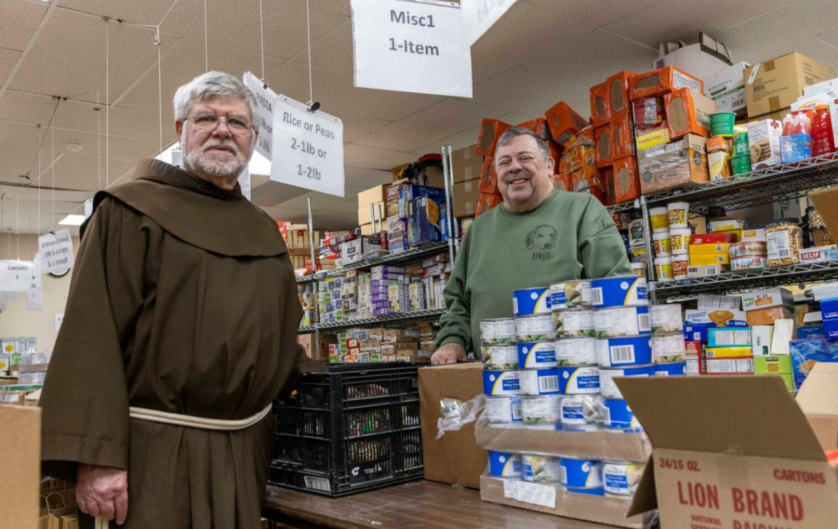 A friar in a brown habit and a man in a green shirt smile as they stand among the shelves of a food pantry