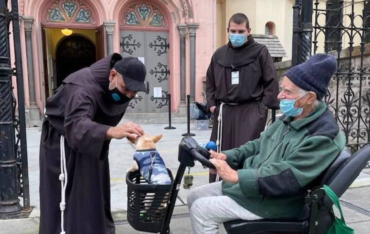 Two friars wearing masks bless a small chihuahua that is sitting in the basket of a mobility scooter driven by an older man wearing a mask