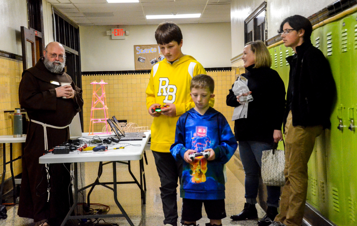 A friar oversees two students using electronics as their relatives look on.