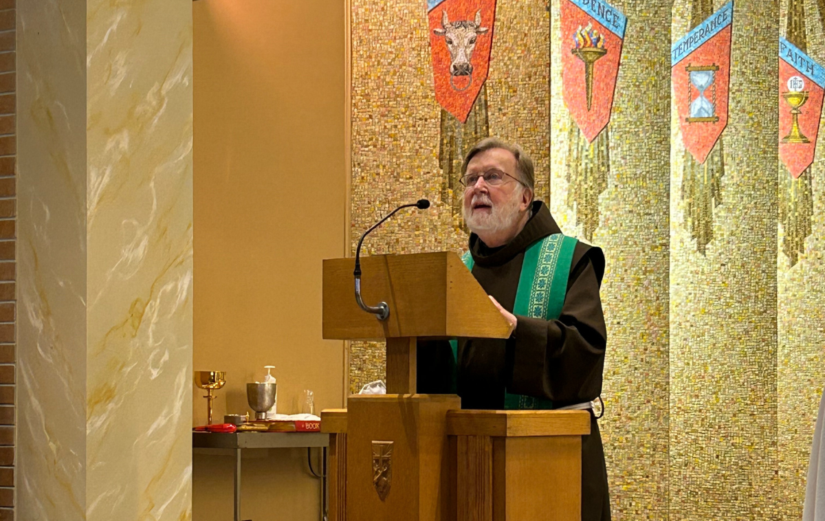 A friar wearing a green stole over his brown habit speak from a lectern.