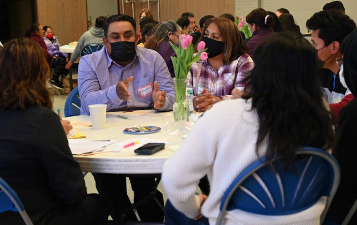 Six people sit at a round table. A man wearing a lavender shirt and a black K-95 mask gestures with his hands as he speaks.