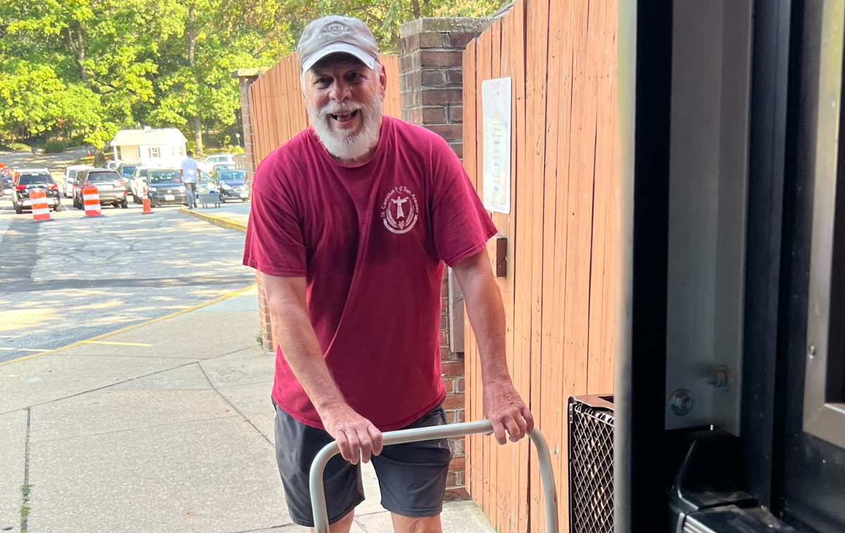 A smiling man wearing a red shirt and grey baseball hat pushes a hand cart.