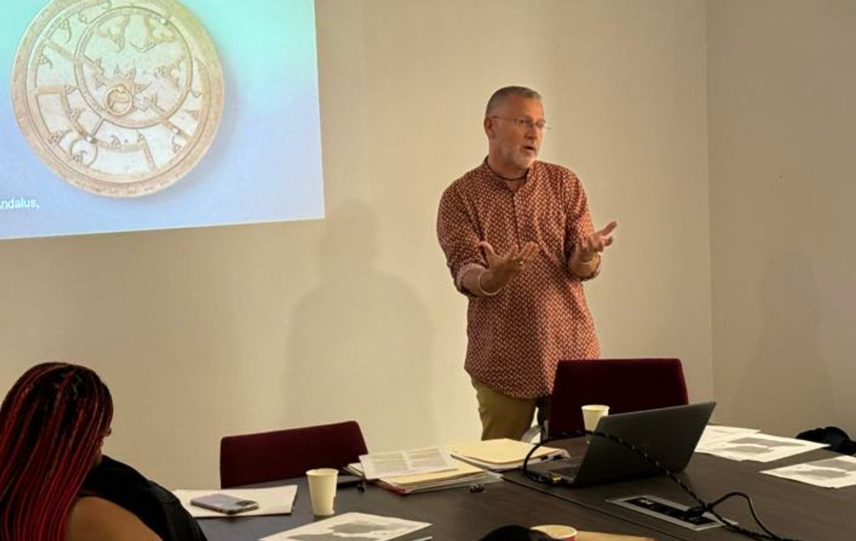 A man wearing a red and white checkered shirt teaches in front of a classroom of adult students.