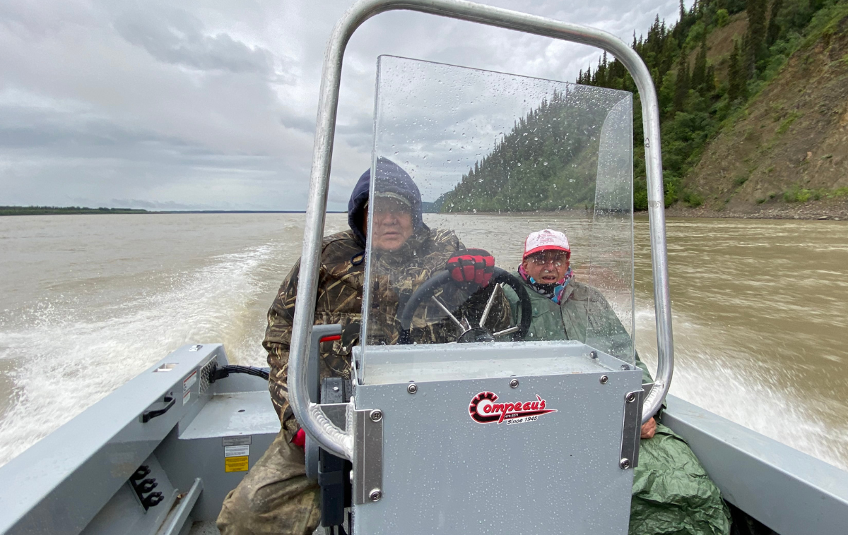 Two men wearing full-length rain jackets and hoods navigate a boat down the Yukon River.