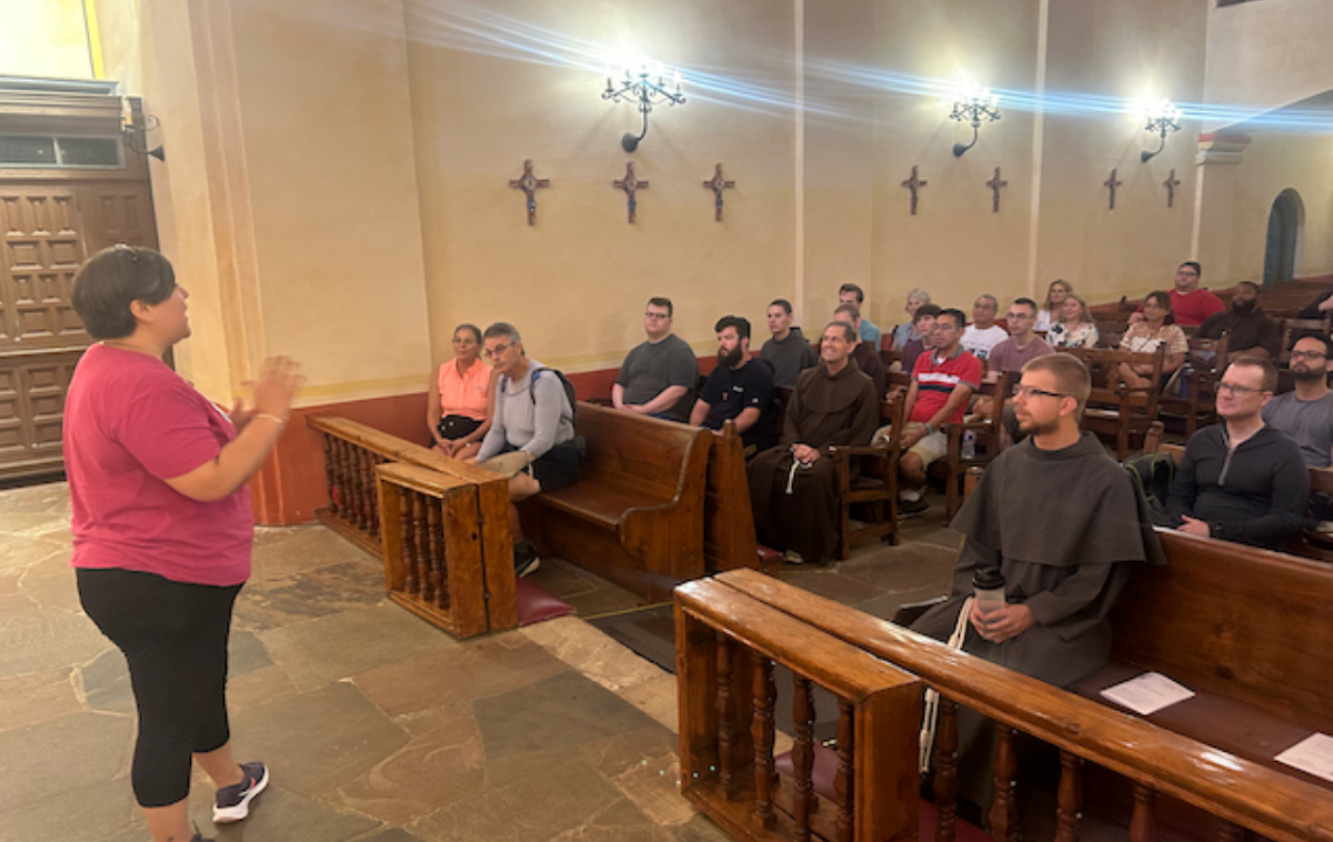 A group of friars and men in discernment sit in the pews of an old mission church and listen to the woman standing at the front of the room.