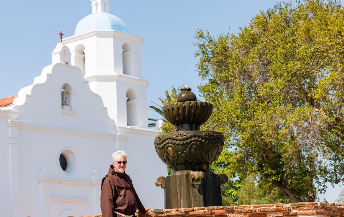 A bearded man with sunglasses and wearing a friar habit stands next to a fountain in front of a white mission-style church.