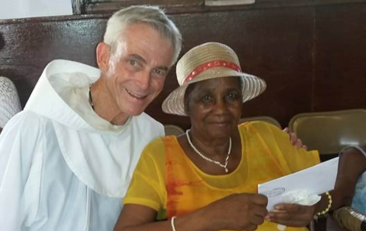 Br. Jim sits next to a parishioner who is wearing a straw hat and a bright yellow blouse. They both smile at the camera.