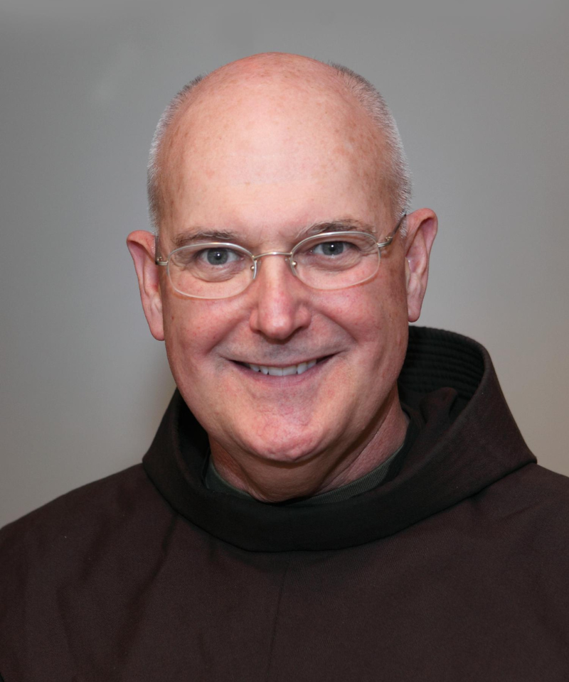 A smiling man in glasses and a friar's robe smiles for a head shot.