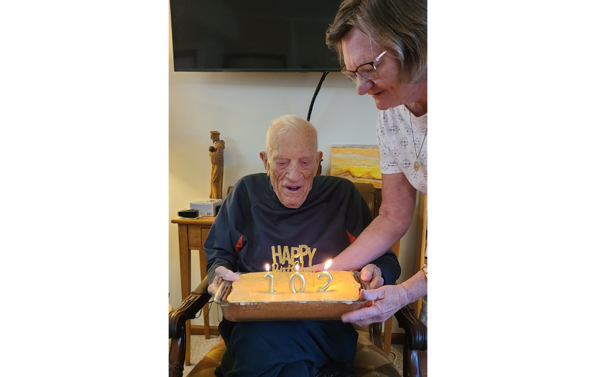 A man seated in a chair is presented a birthday cake by a woman standing next to him.