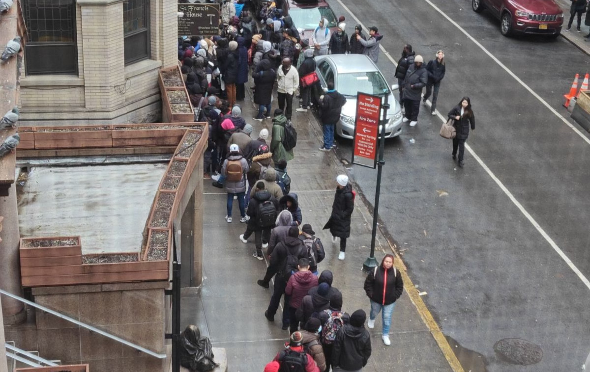 Dozens of people in winter clothing wait in line on a city sidewalk to get into a building.