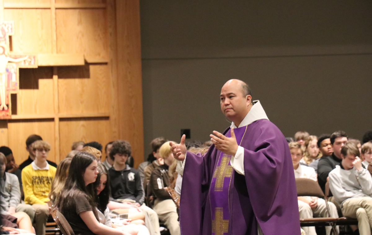 A priest in a purple robe speaks to several seated students.