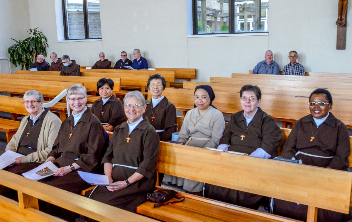 A group of eight Poor Clare Sisters wearing their brown habits sit in the wooden pews of a church. They are smiling at the camera.