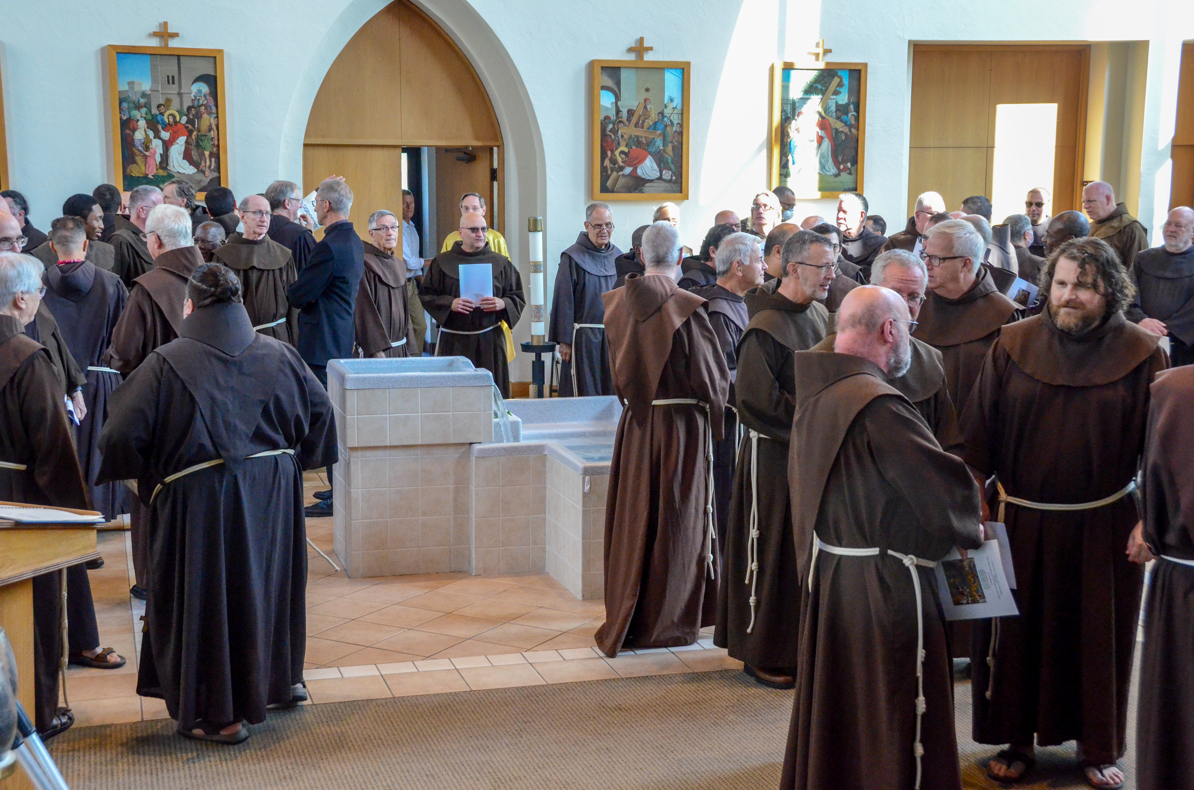 A large group of friars stand in the vestibule of a church in a scattered group, talking amongst each other.