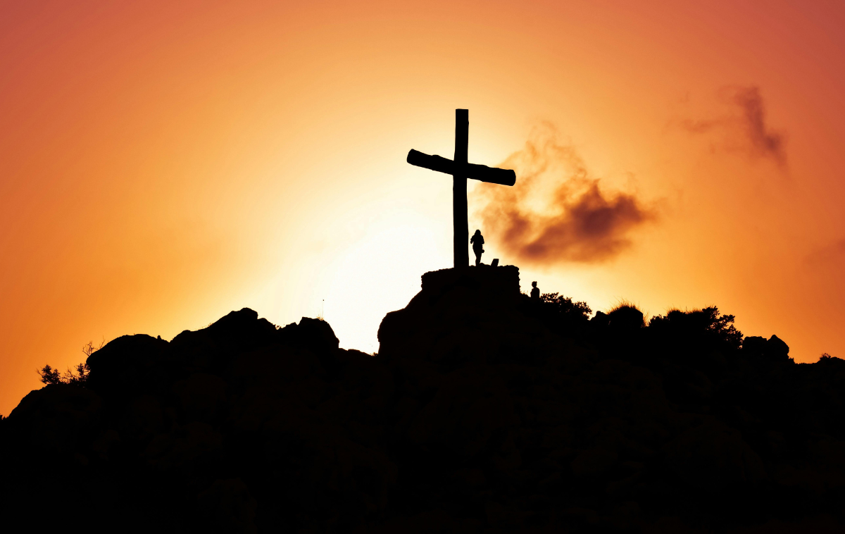 A person stands next to the silhouette of a cross as the sun sets.