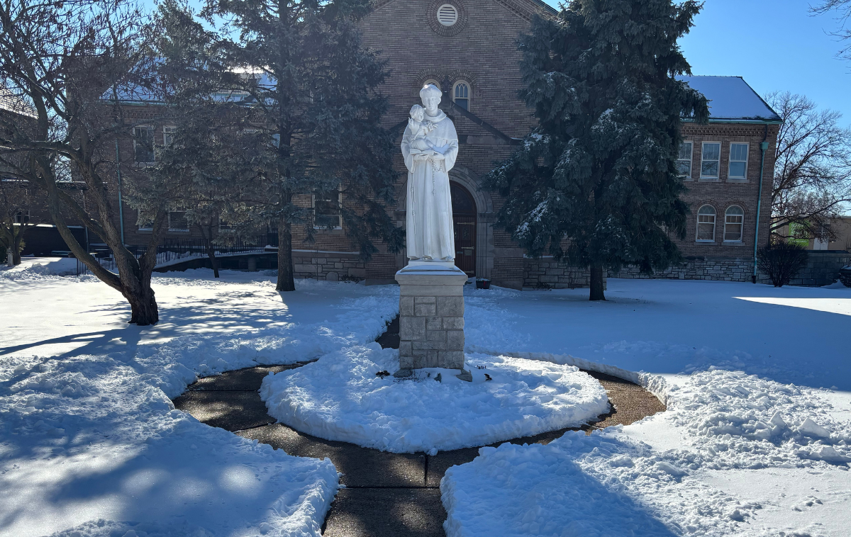 A statue of St. Francis sits in the middle of a rotunda with snow on the ground. There is a building and trees behind it.