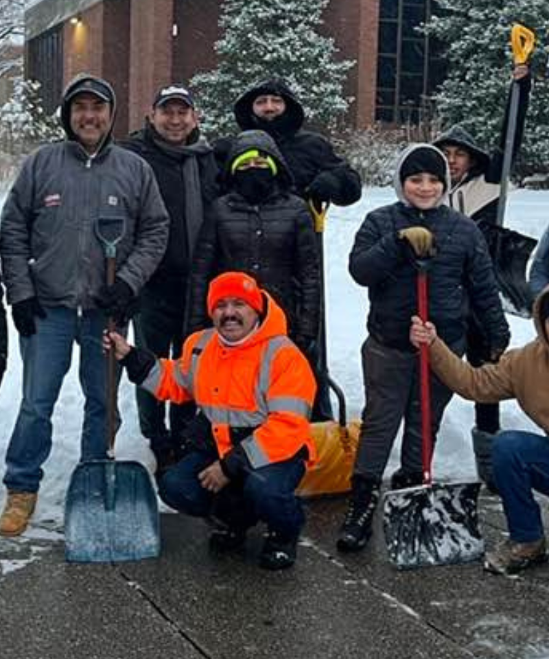 Several smiling people pose for a photo on a sidewalk. They are wearing winter clothing and there is snow on the ground behind them.