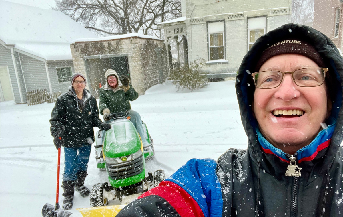 Three people smile for a photo on a snowy street. All are wearing winter clothing and one is sitting on a tractor.