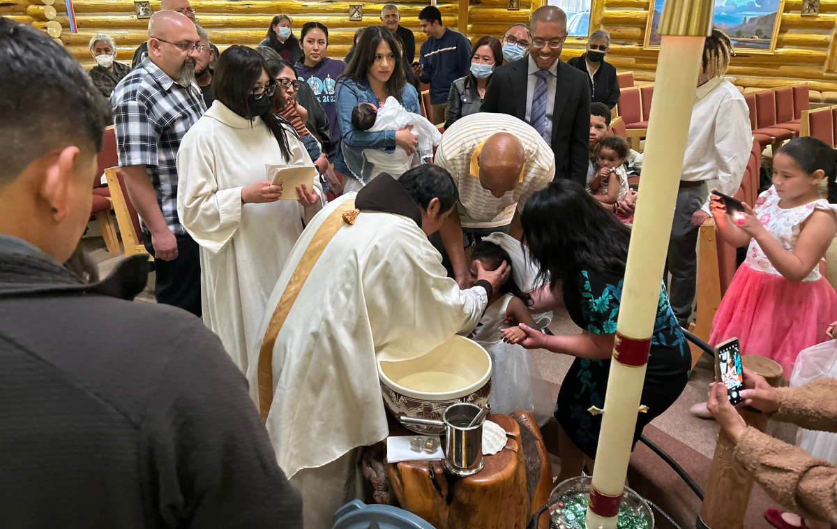 A friar baptizes a baby as a crowd watches.