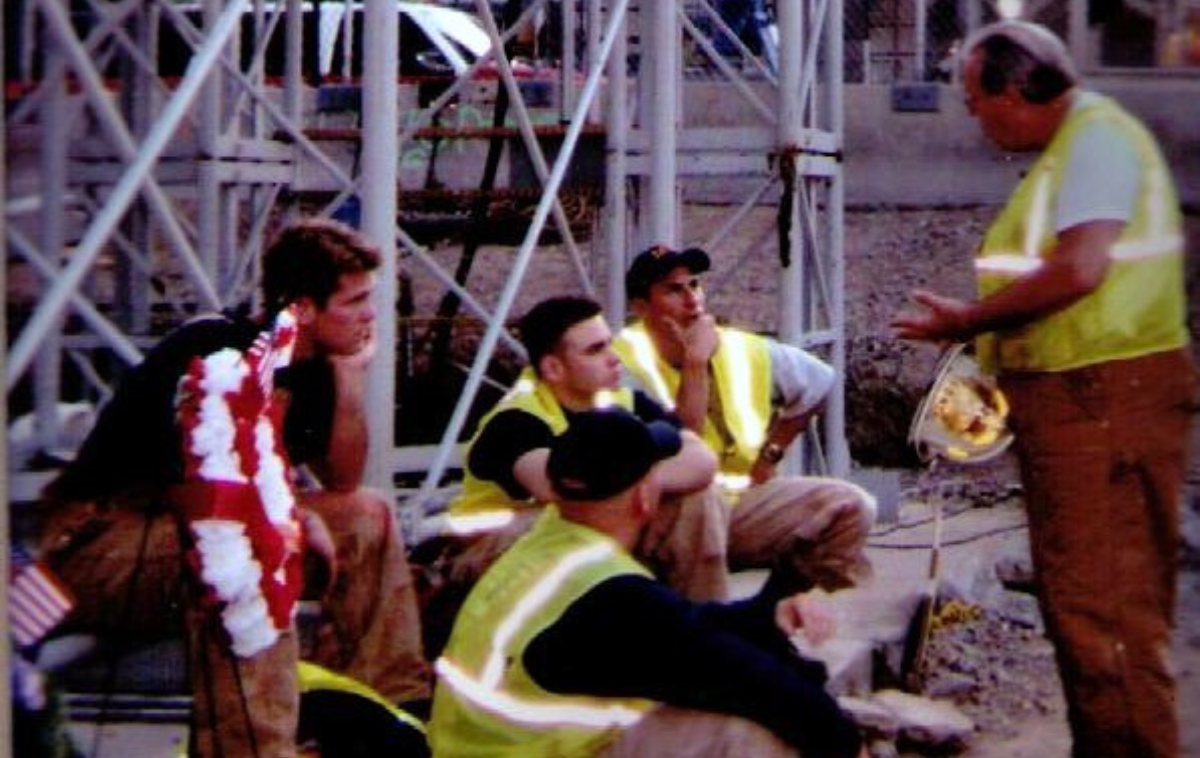 A friar priest stands before a group of seated men. Four of the five are wearing reflective safety vests.