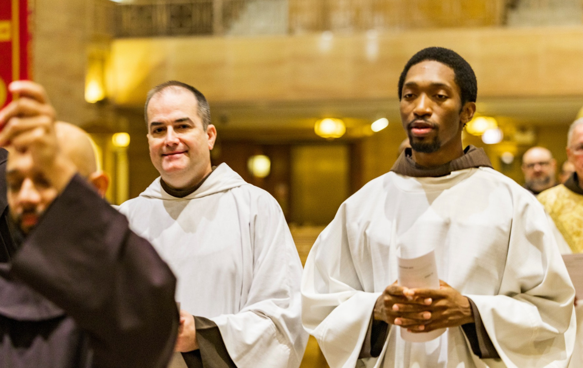 Two men in vestments walk side by side down the center aisle of a Catholic church.