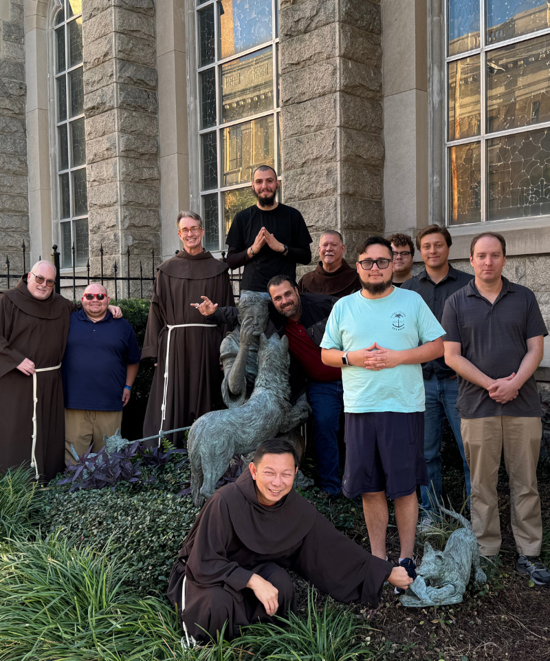 Eleven men pose outside for a photograph around a statue. Four men are wearing friar habits.