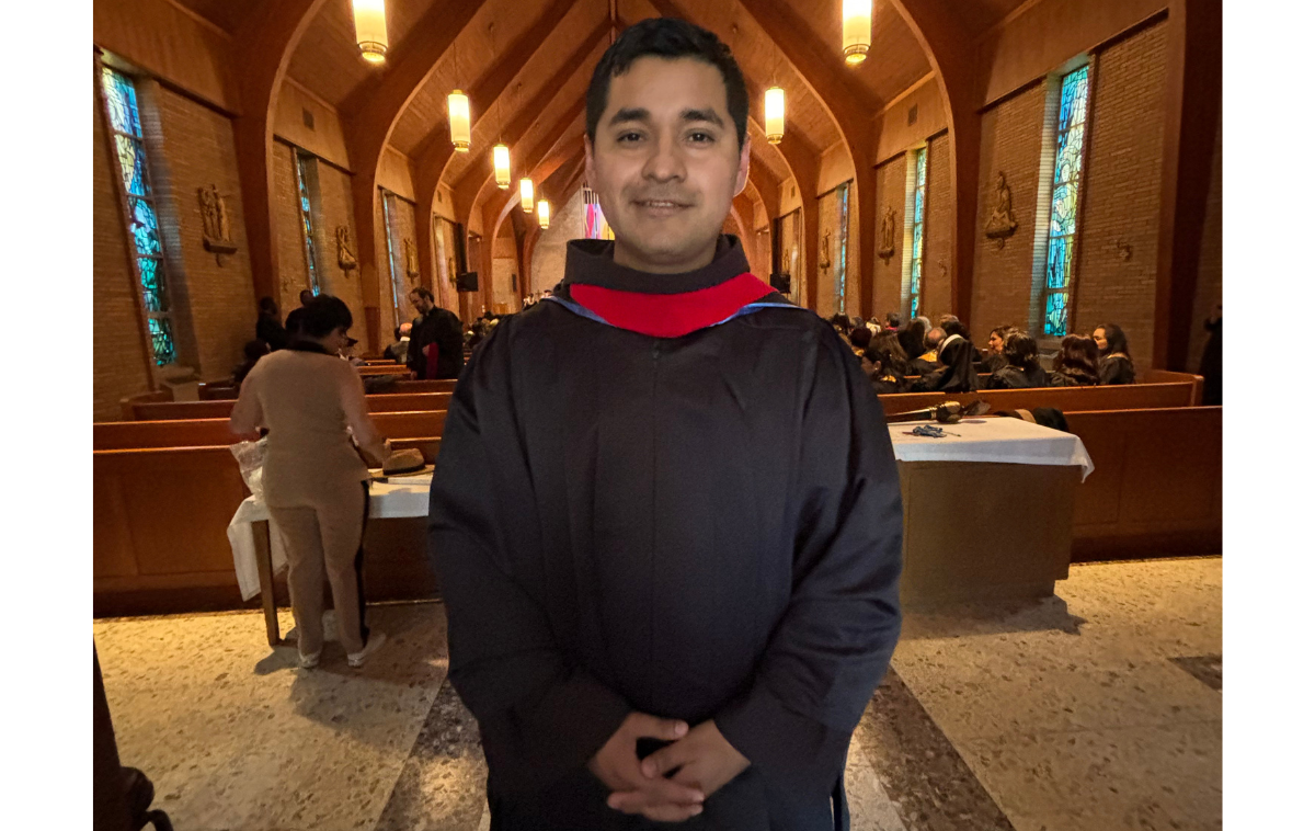 A smiling man stands at the back of a church. He is wearing a friar habit and red sash.