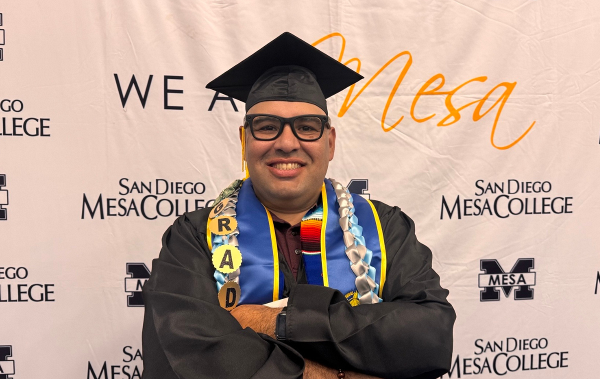 A smiling, bespeckled man poses for a photo wearing a graduation mortar, robe and stole.