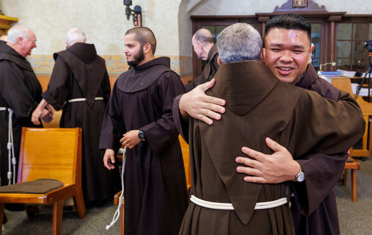 Two men in friars habits embrace. Four more men in friars habits are in the background.