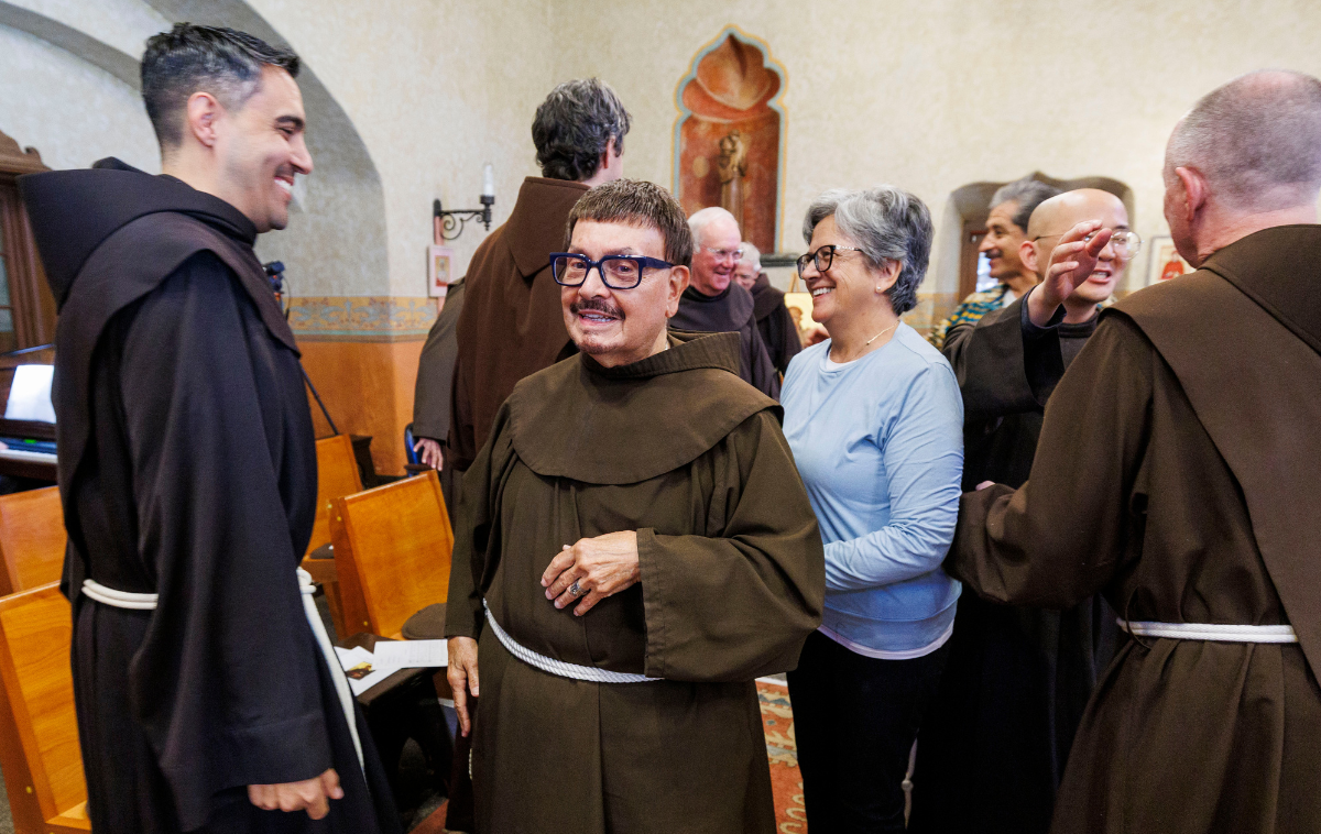 Two smiling friars have a conversation. Friars and a woman in a blue sweater are in the background.