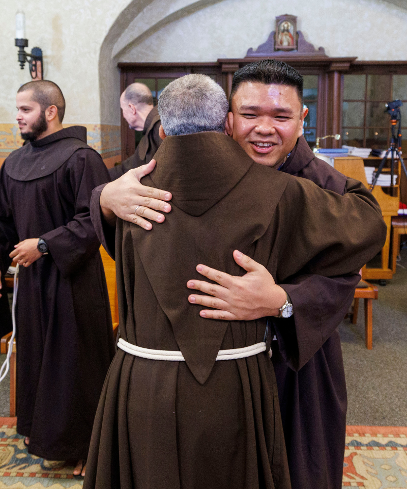 Two men wearing friar habits embrace. Two more friars are in the background.