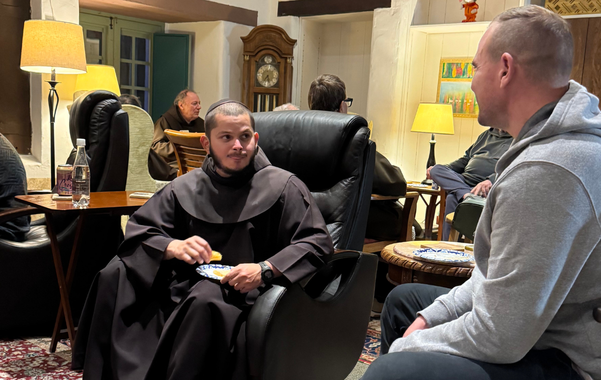 A man in a friar habit sits in a chair and is talking to a man wearing a gray sweatshirt. The friar is holding a plate with food on it.