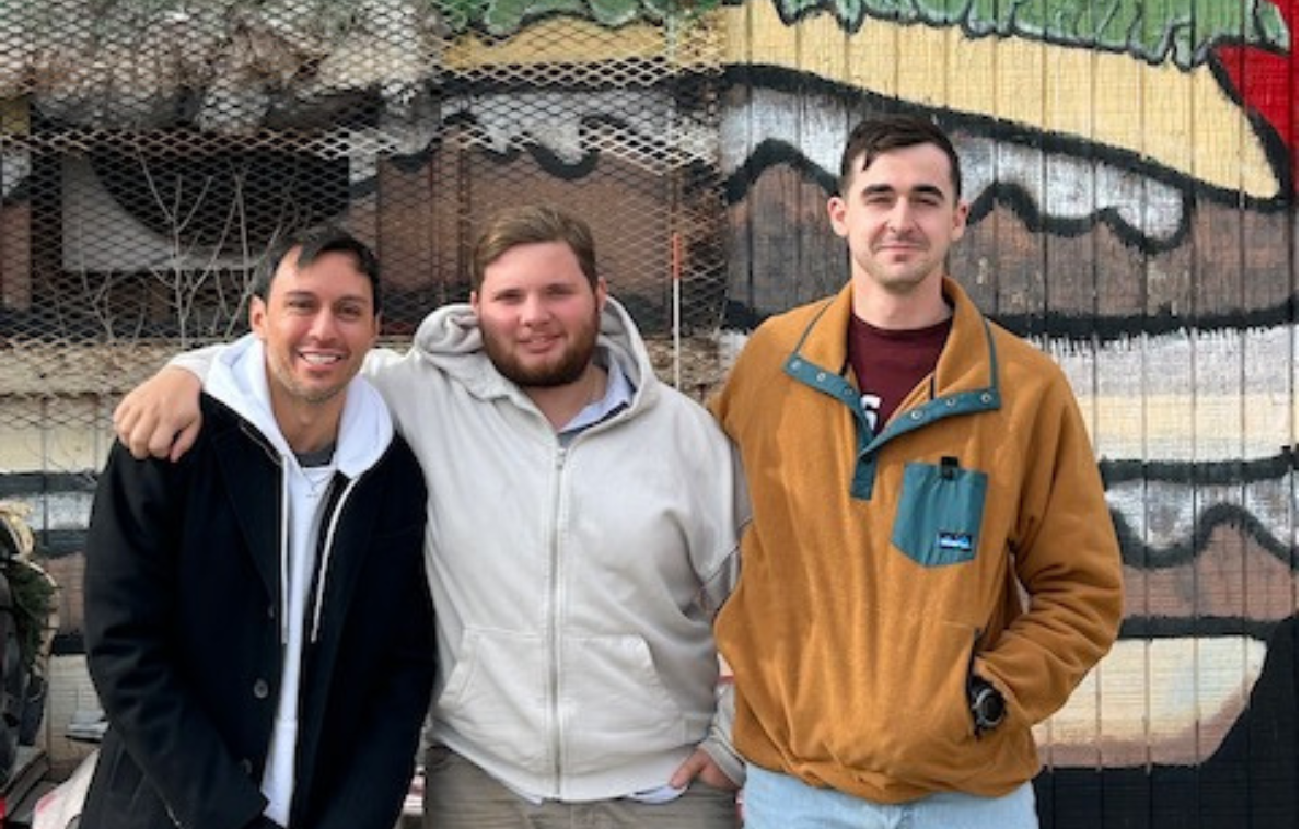 Three smiling men wearing hooded jackets pose for a photograph in front of a wall with a hamburger painted on it.