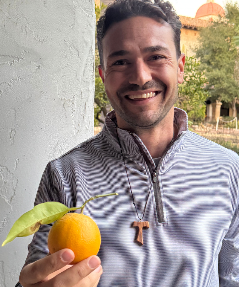 A smiling man wearing a gray jacket and tau cross holds an orange.