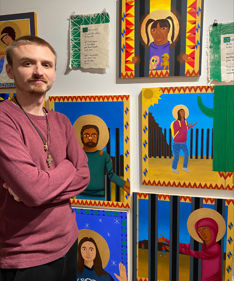 A man wearing a red long-sleeved t-shirt and necklaces stands in front of a wall with six paintings hanging on it.