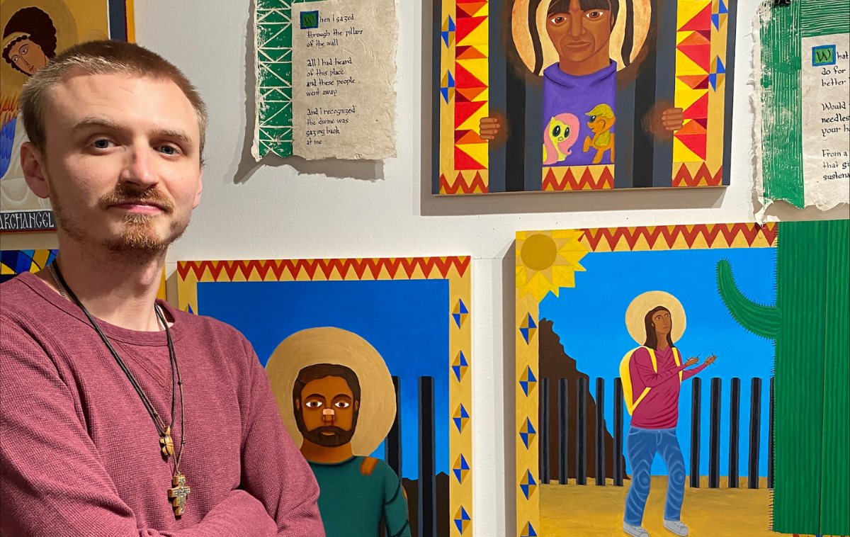 A mean wearing necklaces and a red long-sleeved shirt stands next to a wall with several paintings on it.