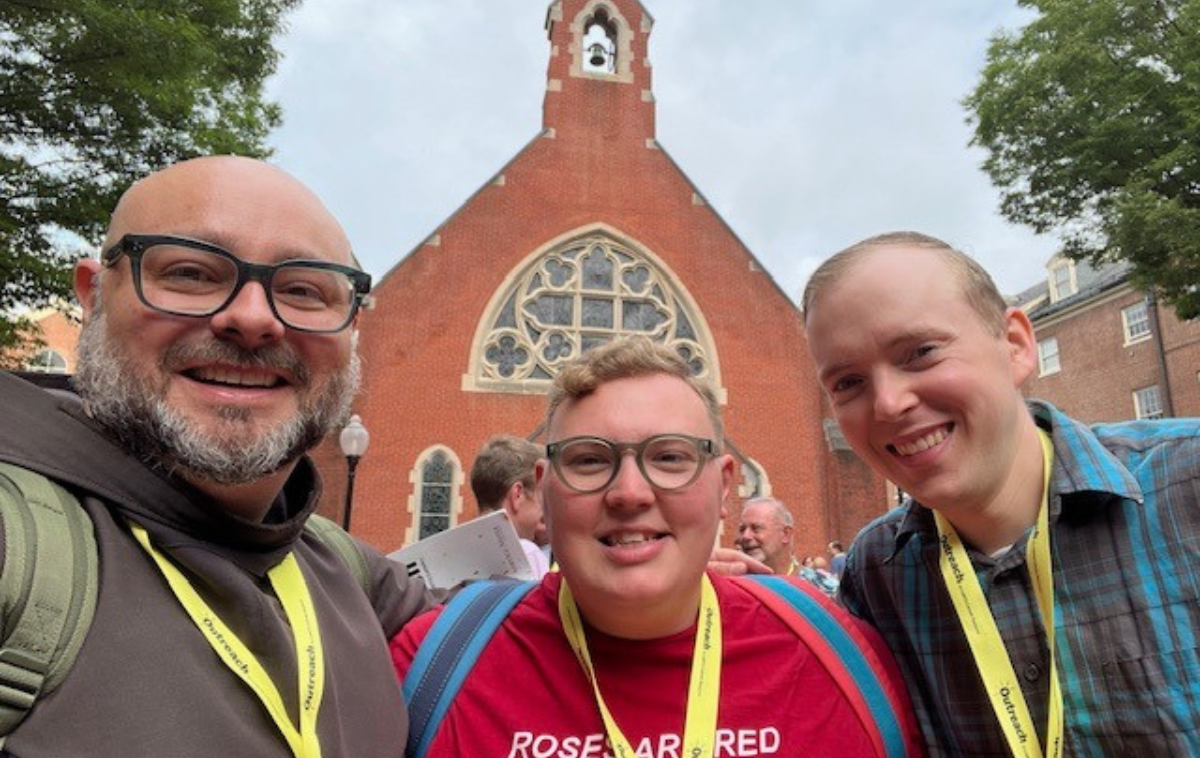 Two bespeckled people and one without glasses pose for a photo. One is bearded and wearing a friar habit. All three are wearing yellow lanyards.