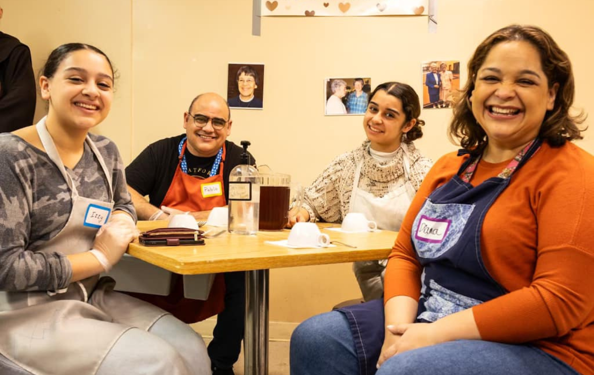 Four smiling people wearing aprons sit at a table. There are masks, a wallet, and two drink pitchers on the table, and three photos on the wall behind them.