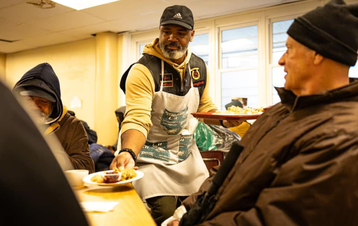 A bearded man wearing a ball cap and apron serves food on plates to men sitting at a table. The man in the foreground is wearing a black knit cap and a brown coat.