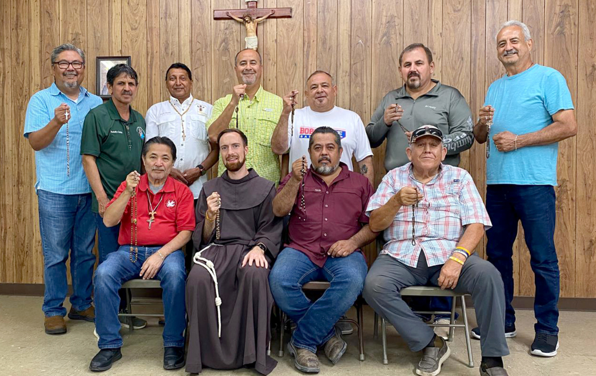 Four seated men, including one wearing a friar habit, and seven standing men pose for a photo, some holding up rosaries.