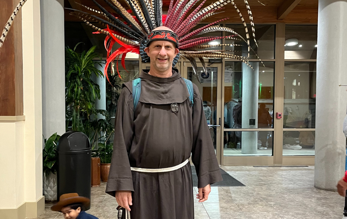 A man wearing a friar habit wears a headdress of red and yellow feathers. A child in the foreground is wearing a brown hat and blue shirt.