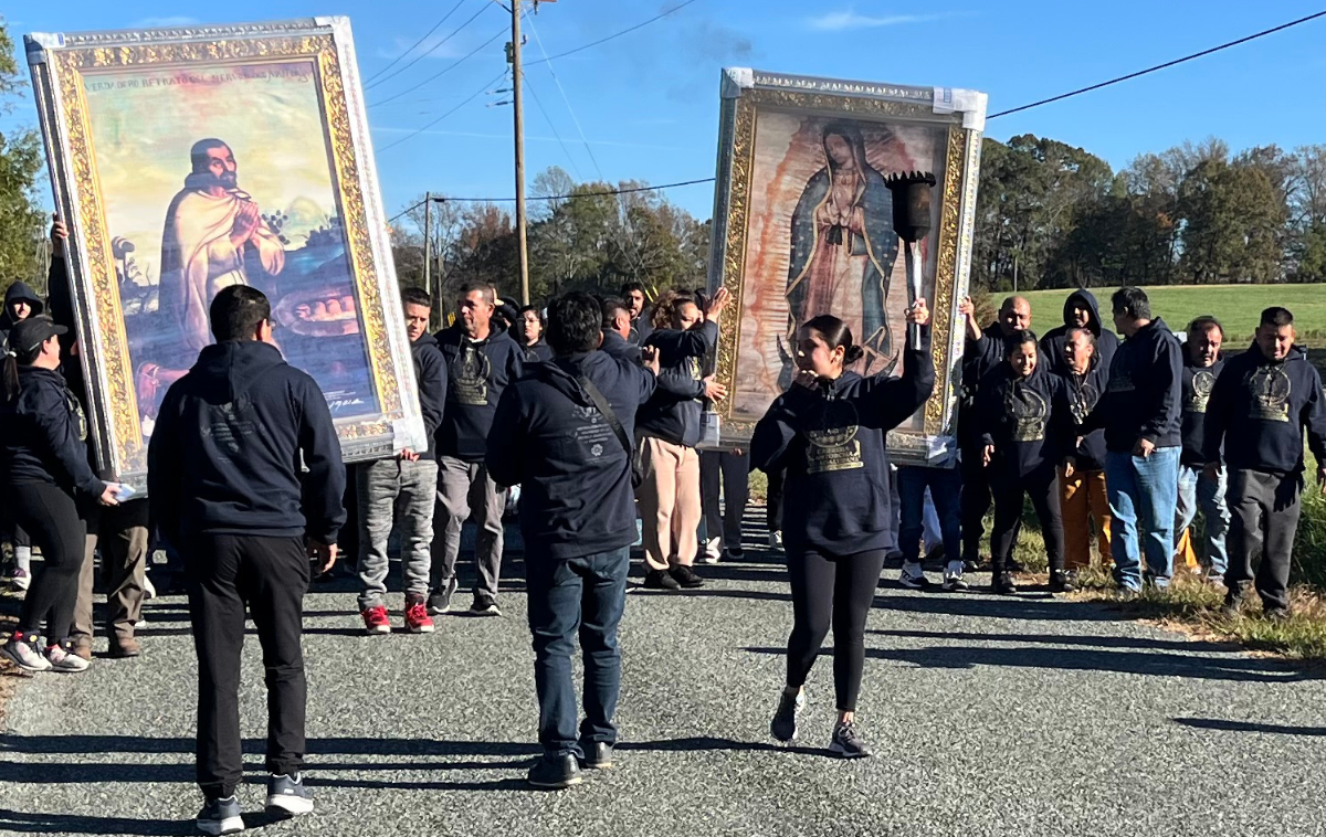 Several people walk on a road wearing blue hooded sweatshirts. Some are carrying frames with religious images on them, and one woman is holding a torch.