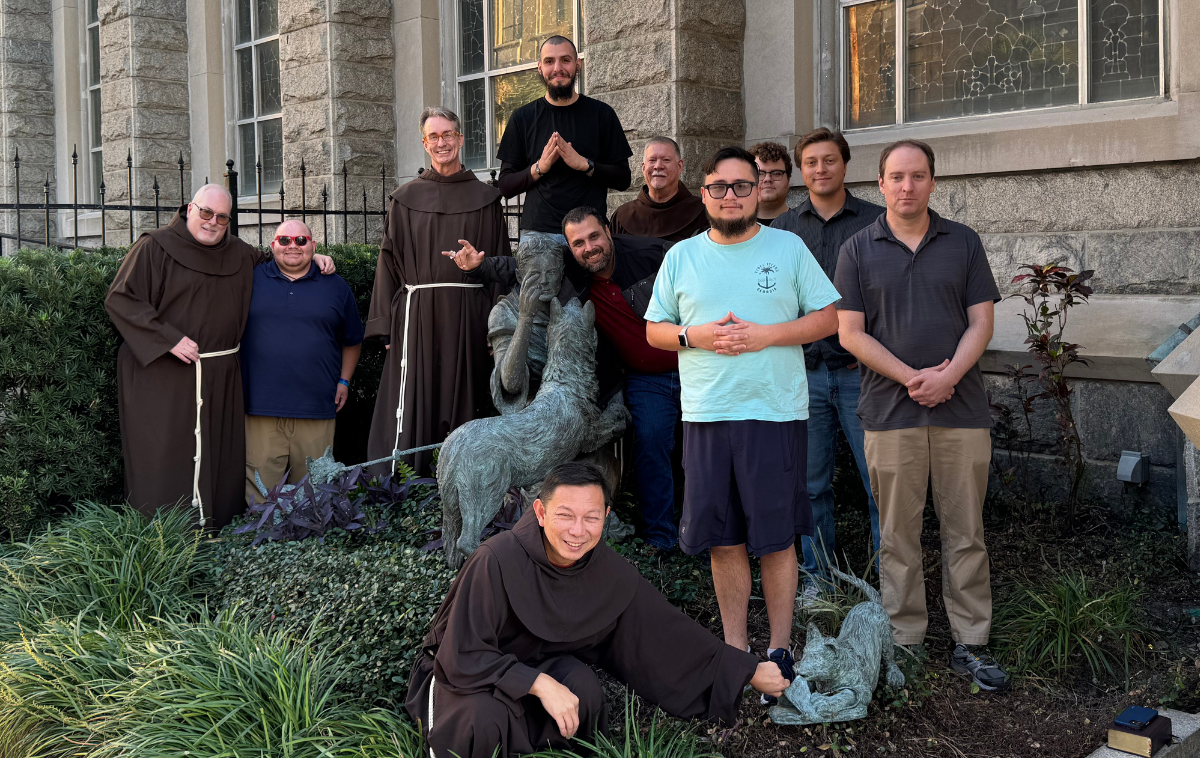 Eleven men pose outdoors for a photo with a statue. Four are wearing friar habits.