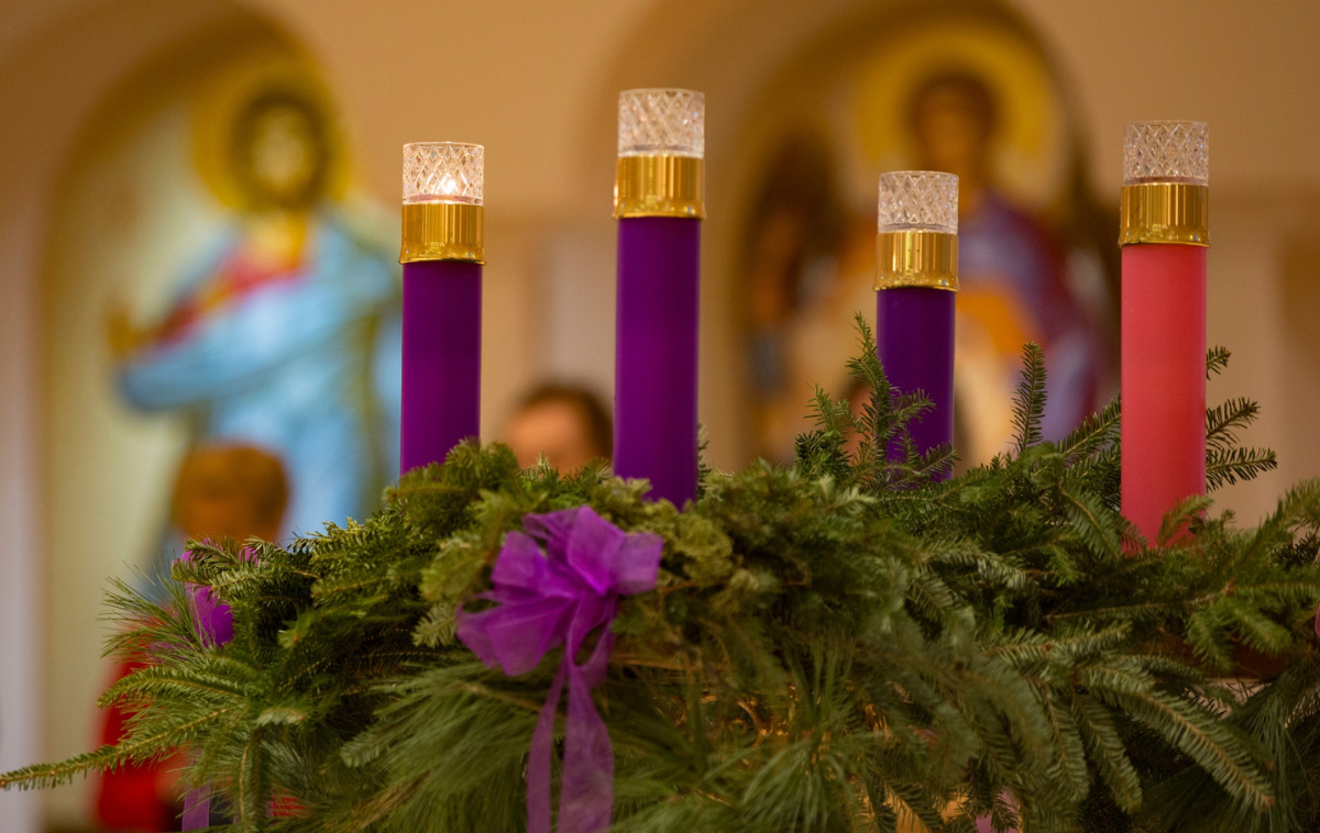 An advent wreath with three purple candles and one pink one sit in a church.