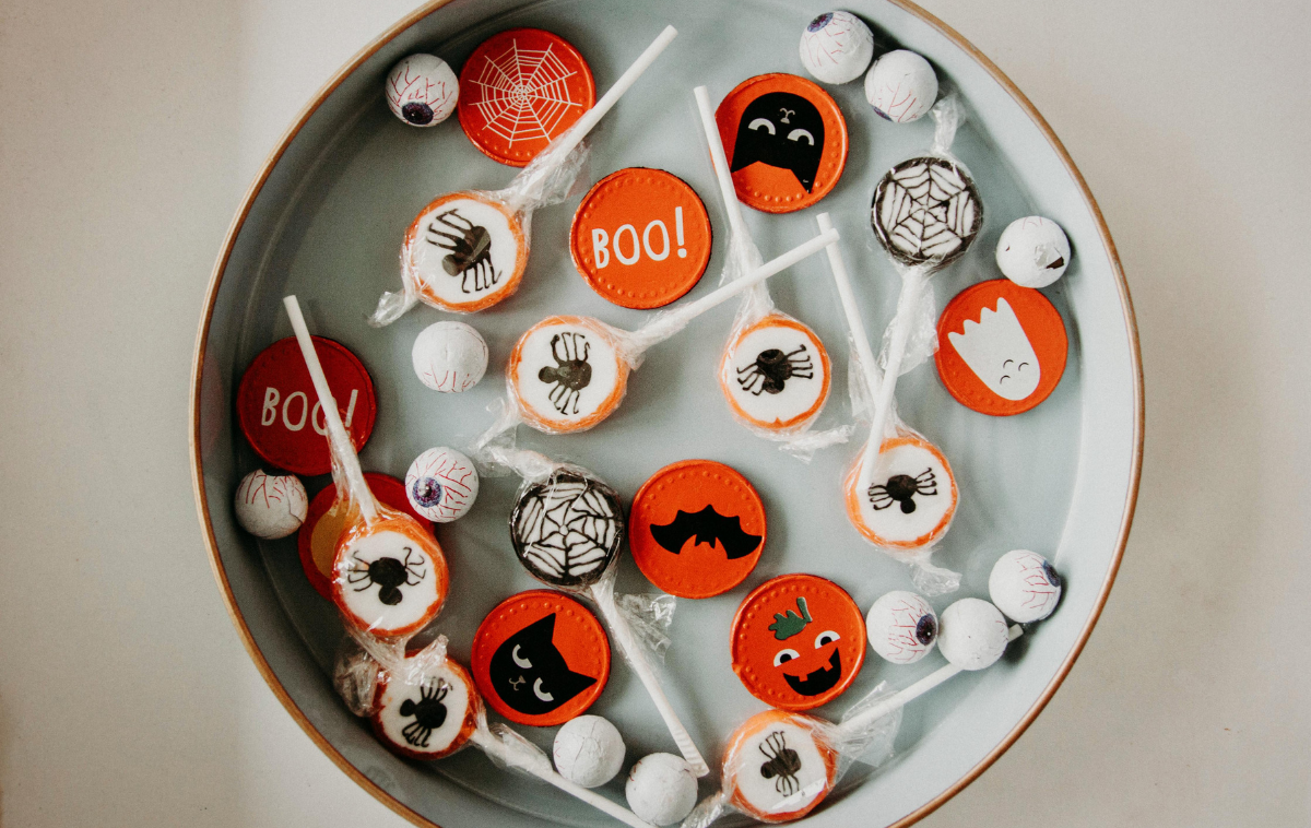 A dish full of Halloween candy, including lollipops with spider images on them, candies wrapped in orange foil featuring images of bats, cats, and jack-o-lanterns, and round candies that look like eyeballs.